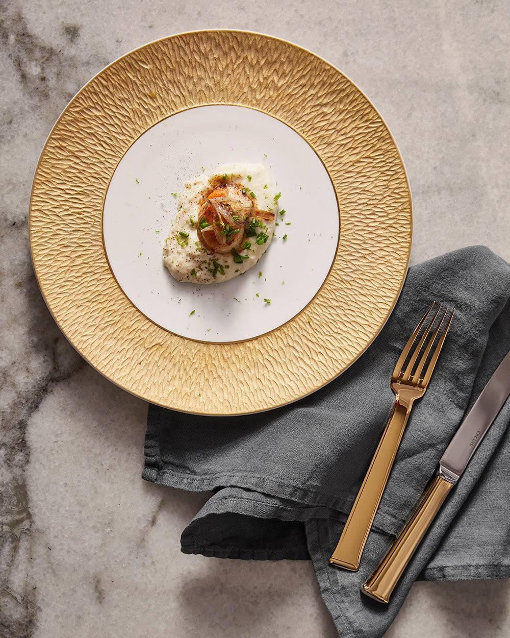 Gold-rimmed textured plate with gold Ercuis cutlery on a marble table