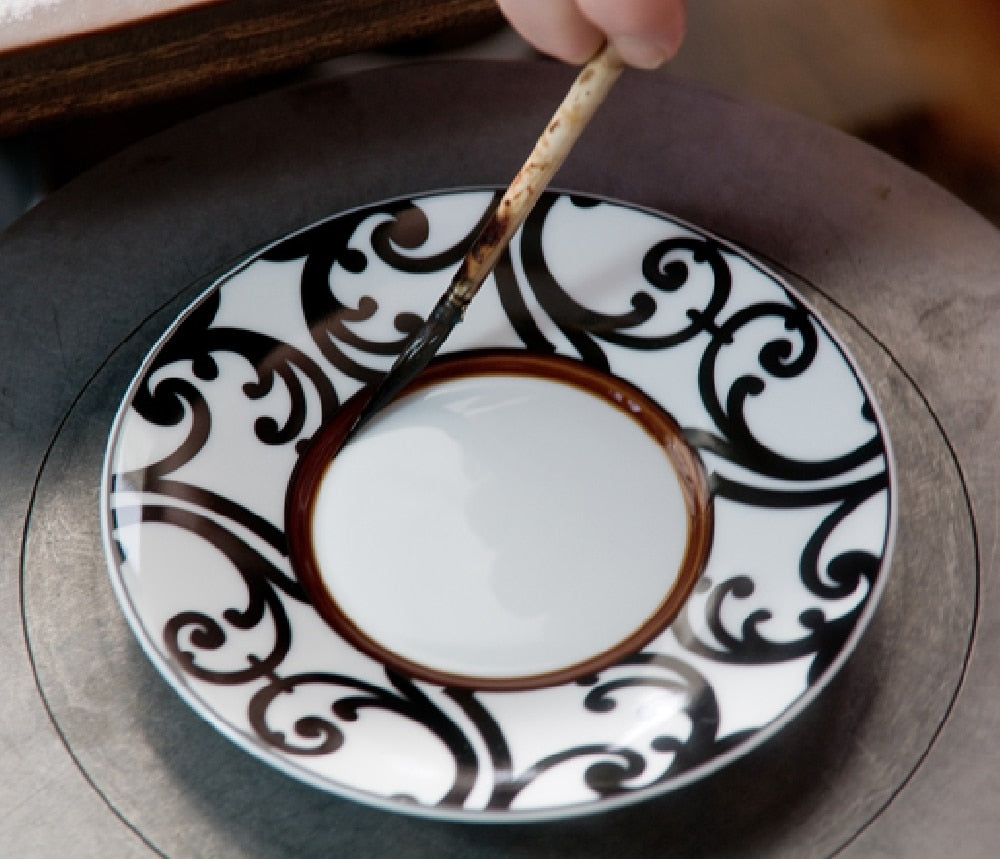 Black scrolls being painted by hand on a Seignolles porcelain saucer