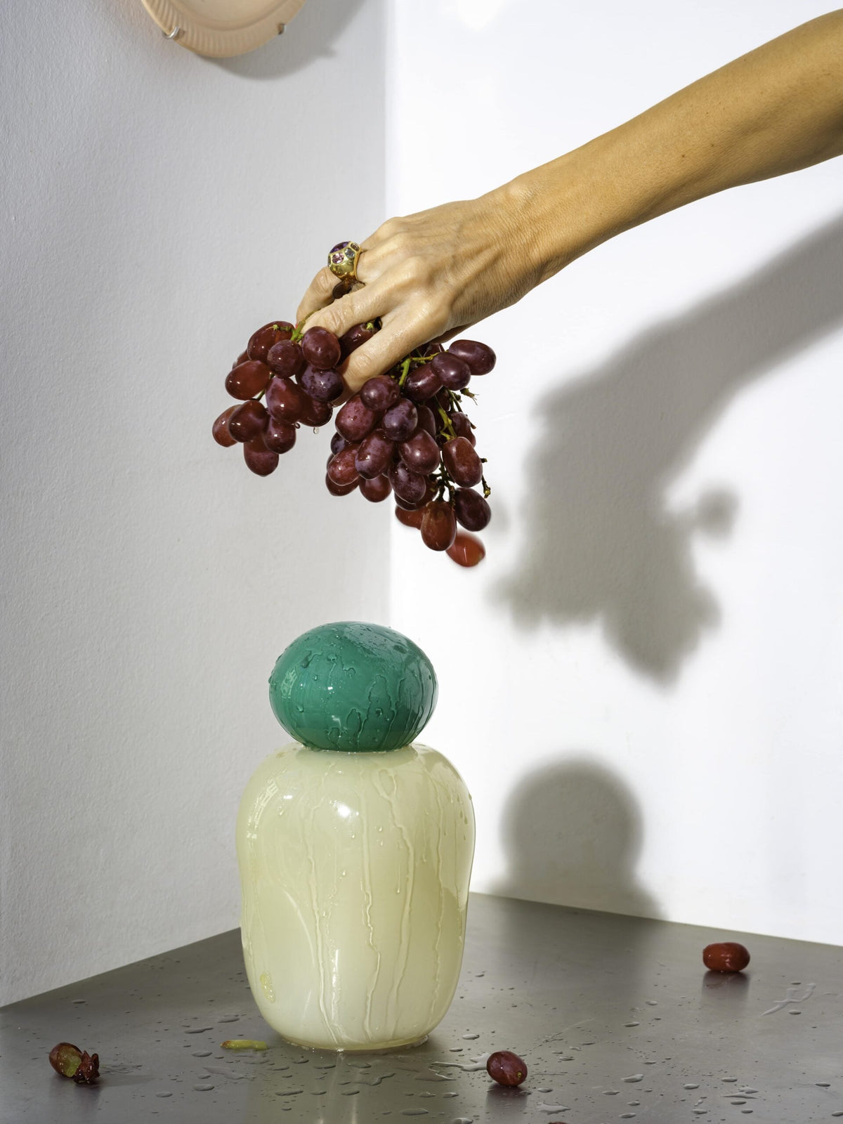 A hand holding grapes above a Helle Mardahl mouth-blown glass jar in cream and green