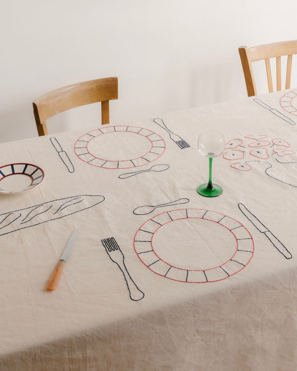 A linen tablecloth embroidered by Sarah Espeute with outlines of plates, cutlery and bread, beside wooden chairs