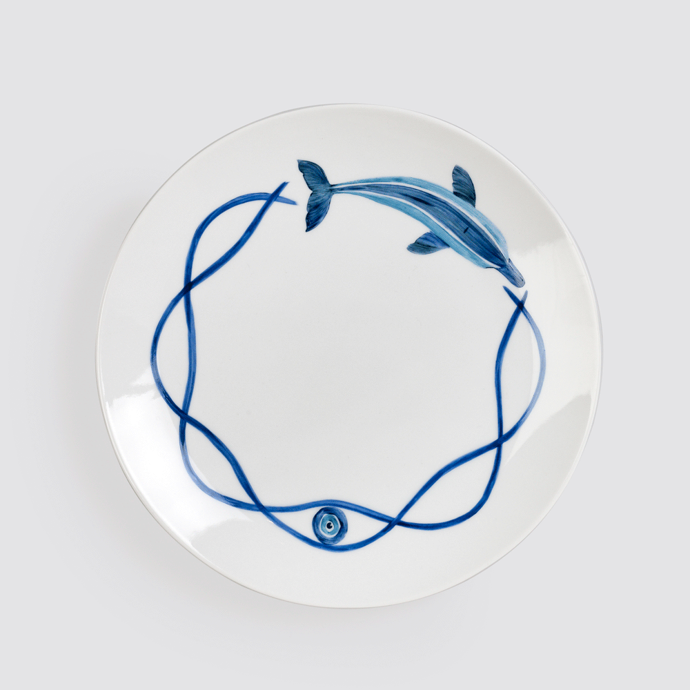A white plate from Victoria de Borbón's Dolphin Collection, painted in blue with a dolphin and an evil-eye motif