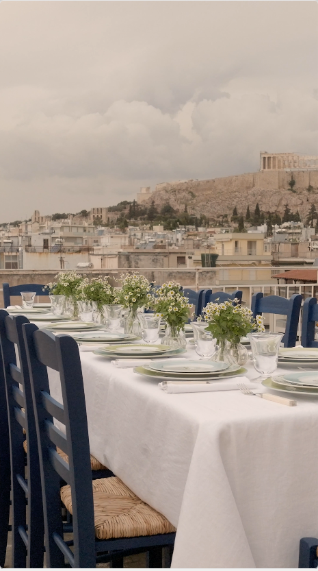 A long table set with Victoria de Borbón's Greek Collection on an Athens rooftop, with the Acropolis behind